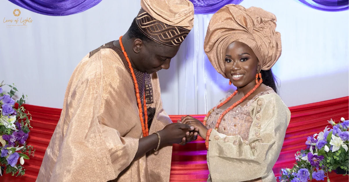 A Nigerian couple in traditional gold and brown Aso Oke attire, with the groom holding the bride's hand during a wedding ceremony.