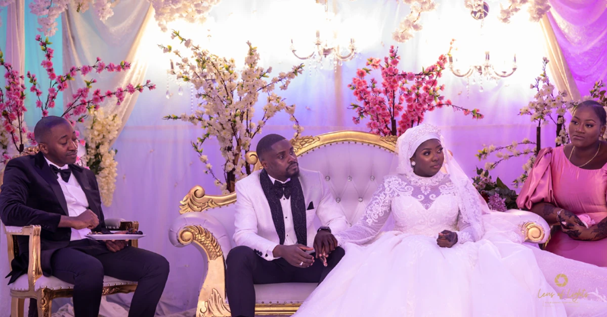 A bride and groom sitting on a gold-trimmed white settee during a UK wedding reception, surrounded by pink and white floral decor.