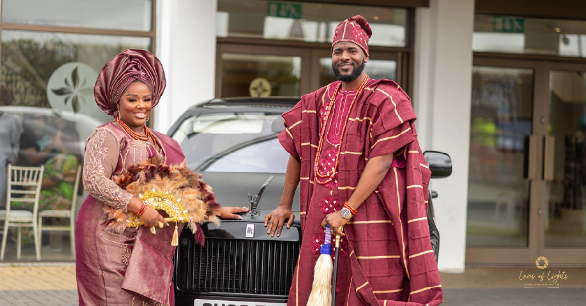 A Nigerian couple in traditional maroon Igbo wedding attire standing in front of a black Rolls Royce in the UK.